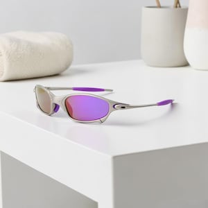 May include: Silver-framed sunglasses with purple lenses and accents. The sunglasses have a sleek, modern design with a curved frame and a pop of purple on the temples. The lenses reflect a gradient of colors.