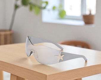 May include: Gray shield sunglasses with a gold star accent on the temple. The sunglasses have a mirrored lens and are resting on a light brown wooden table. The overall style is modern and futuristic.