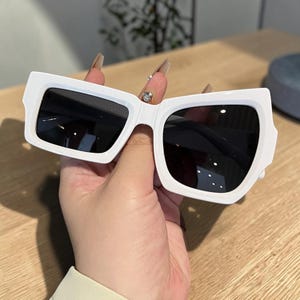 May include: White sunglasses with a thick, rectangular frame and dark lenses. The sunglasses have a modern, angular design. The frame is a bright white colour, and the lenses are black. The sunglasses are held in a hand.