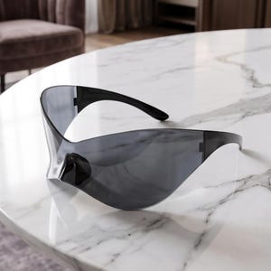 Puede incluir: Unas gafas de sol tipo escudo futuristas con una lente única continua de color gris oscuro que envuelve la parte frontal. La montura está fabricada en plástico negro con patillas elegantes y curvadas que salen de los lados de la lente.