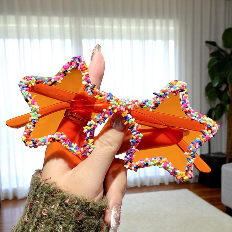 K&ouml;nnte beinhalten: Orange sternf&ouml;rmige Sonnenbrille mit buntem Streuselrand. Die Sonnenbrille hat orangefarbene Gl&auml;ser und B&uuml;gel. Der Rahmen ist mit einer lebendigen Anordnung von Streuseln verziert, die der Brille eine verspielte und einzigartige Note verleihen.