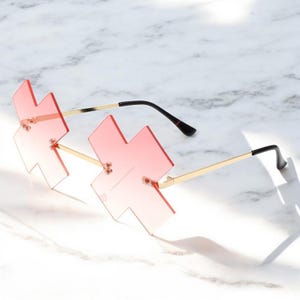 Könnte beinhalten: Eine Sonnenbrille in Pink und Gold mit einem Kreuzrahmen. Die Sonnenbrille hat ein Verlaufsglas, das von einem dunkleren Pink zu einem helleren Pink übergeht.