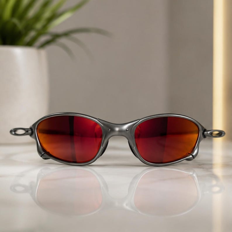 Peut inclure: Lunettes de soleil avec une monture gris m&eacute;tal et des verres ovales dot&eacute;s d'une teinte rouge-orange vive et r&eacute;fl&eacute;chissante. La monture pr&eacute;sente un pont sculpt&eacute; distinctif et des branches fines et incurv&eacute;es sur les c&ocirc;t&eacute;s.