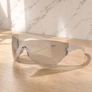 Op de afbeelding: Transparante, oversized zonnebril met een wrap-around design. De lenzen zijn lichtgrijs getint en het montuur is van transparant plastic. De zonnebril heeft een moderne, futuristische uitstraling.