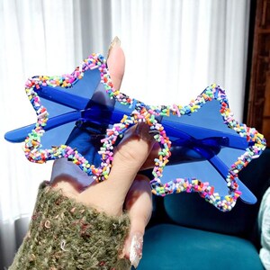 K&ouml;nnte beinhalten: Blaue sternf&ouml;rmige Sonnenbrille mit einem bunten, streuselartigen Rand. Die Sonnenbrille hat ein klassisches Rahmendesign. Der Rahmen hat eine leuchtend blaue Farbe. Sie wird von einer Hand gehalten.