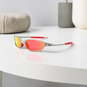 May include: Silver-framed sunglasses with red and orange lenses. The temples and nose bridge have red accents. The sunglasses are on a white surface.