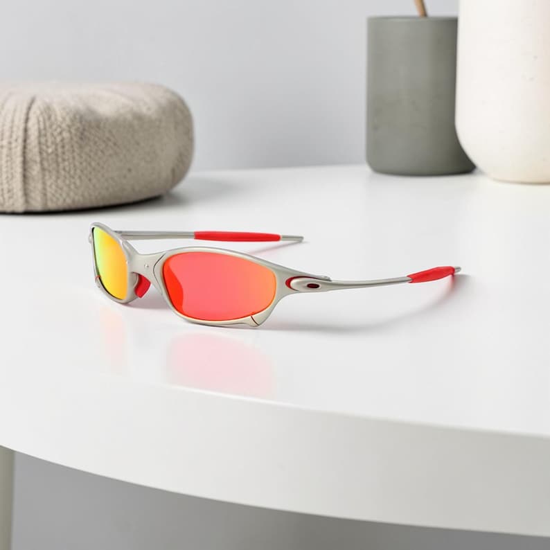 May include: Silver-framed sunglasses with red and orange lenses. The temples and nose bridge have red accents. The sunglasses are on a white surface.