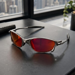 Peut inclure: Une paire de lunettes de soleil dot&eacute;e d'une monture en m&eacute;tal argent&eacute; et de verres rouges vifs. Ces lunettes pr&eacute;sentent un design enveloppant avec des branches fines et profil&eacute;es aux extr&eacute;mit&eacute;s sombres, affichant une structure industrielle.