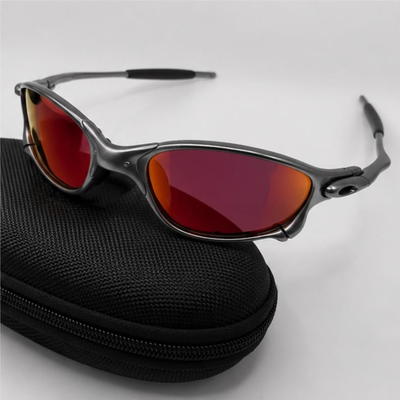 Peut inclure: Lunettes de soleil avec une monture gris acier m&eacute;tallis&eacute; et des verres rouges r&eacute;fl&eacute;chissants. Les lunettes pr&eacute;sentent un design enveloppant avec des embouts de branches noirs et sont pos&eacute;es sur un &eacute;tui de transport rigide noir textur&eacute;.
