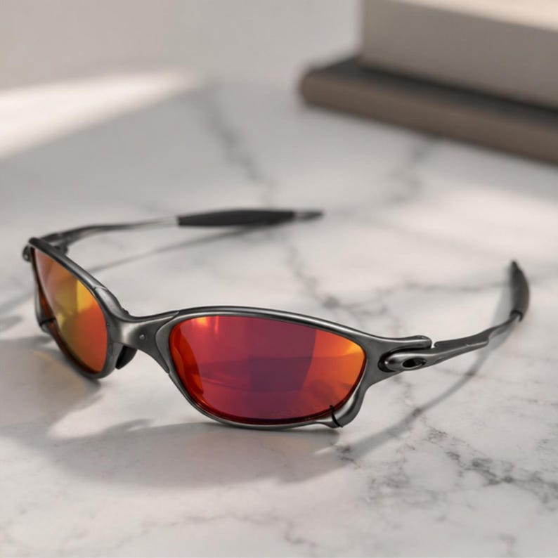 Peut inclure: Lunettes de soleil avec une monture gris m&eacute;tallique fonc&eacute; et des verres r&eacute;fl&eacute;chissants rouge orang&eacute;. Les lunettes pr&eacute;sentent un design enveloppant avec des branches fines et incurv&eacute;es et un pont reliant les deux verres ovales.