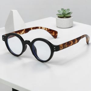 Könnte beinhalten: Brille mit schwarzem Rahmen, runden Gläsern und Bügeln im Schildpattmuster. Die Brille liegt auf einer weißen Oberfläche. Im Hintergrund ist eine kleine Sukkulentenpflanze in einem weißen Topf zu sehen.