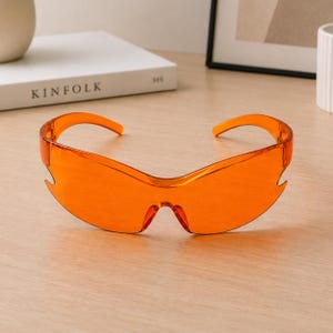 May include: Wraparound sunglasses featuring a single-piece, translucent orange lens and matching orange plastic frames. The design includes a sharp, angular cutout detail on the outer edges of the lens, providing a continuous, sleek, and aerodynamic profile.