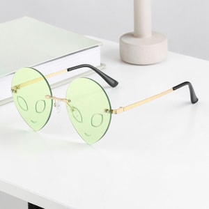 May include: Unique green alien-shaped sunglasses with gold-tone metal frames and black temples. The lenses are a light green color, and the frames are rimless, giving them a modern look. The sunglasses are designed to resemble an alien face.