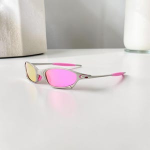 May include: Silver-framed sunglasses with pink and yellow lenses. The temples and temple tips are a bright pink. The sunglasses are on a white surface.