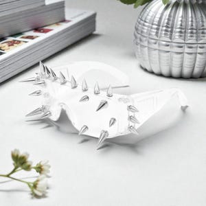 May include: Transparent plastic sunglasses adorned with silver metal spikes. The sunglasses feature a bold, futuristic design, with numerous spikes along the frame. The image is set against a white backdrop, with a decorative vase and a stack of papers in the background.