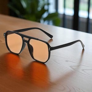 May include: Black square-framed sunglasses with orange-tinted lenses. The sunglasses have a double bridge and are set on a wooden surface. The frames have small silver accents on the corners.