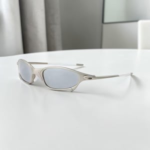 May include: Silver-framed sunglasses with gray lenses. The sunglasses have a sleek, modern design with curved frames and white temple tips. The lenses are reflective, and the overall aesthetic is minimalist and sporty.