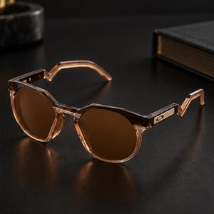 May include: Brown-tinted sunglasses with a translucent dark brown frame featuring a distinct angular zig-zag design on the temples. The frames have a rounded shape with a metallic accent on the side near the hinge, displaying the Oakley logo.