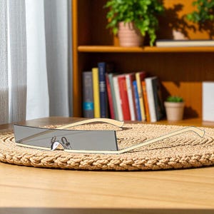 Op de afbeelding: Rechthoekige zonnebril met een lichtgouden montuur en grijze glazen ligt op een ronde, geweven mat. De zonnebril heeft een strak, modern design. De achtergrond toont een houten plank met boeken en planten.