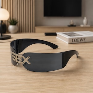 May include: A pair of black wraparound sunglasses featuring a single continuous dark lens and a silver-toned metal accent on the left side displaying the word SEX in a bold, block-style font secured with small visible screws at the temple connection.