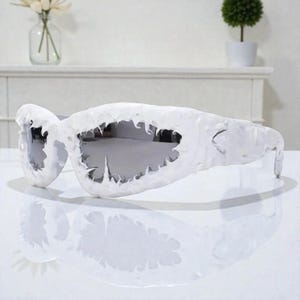 Könnte beinhalten: Weiße Sonnenbrille mit einem einzigartigen, strukturierten Design. Die Fassungen haben eine unregelmäßige, organische Form, und die Gläser sind silberfarben reflektierend. Die Sonnenbrille steht vor einem weißen Hintergrund und erzeugt eine saubere und moderne Ästhetik.