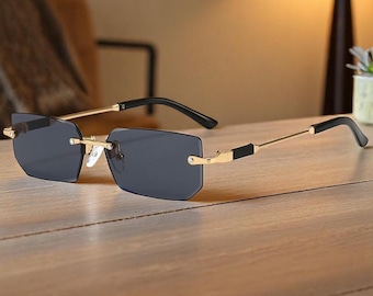 May include: Rimless sunglasses with rectangular, dark gray lenses. The frames are gold-colored with black temples and accents. The sunglasses are displayed on a light brown wooden surface.