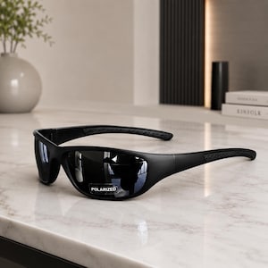 May include: Black sunglasses with a wrap-around frame design and dark, reflective lenses. A small rectangular sticker on the right lens features the white text POLARIZED. The temples have a textured grip pattern along the sides of the frame.
