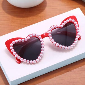 May include: Red heart-shaped sunglasses with dark lenses. The frames are adorned with small, white faux pearls. The sunglasses have a cat-eye shape at the top of the frame.