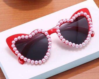 May include: Red heart-shaped sunglasses with dark lenses. The frames are adorned with small, white faux pearls. The sunglasses have a cat-eye shape at the top of the frame.