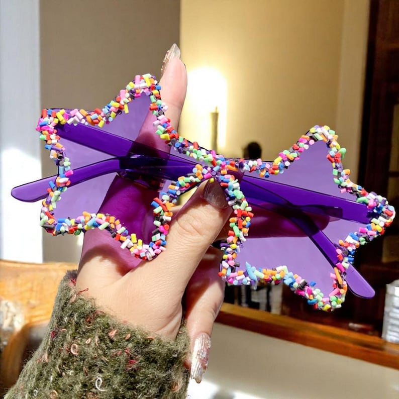 K&ouml;nnte beinhalten: Violette sternf&ouml;rmige Sonnenbrille mit einem bunten, streuselartigen Rand. Die Sonnenbrille hat einen durchgehenden violetten Rahmen und Gl&auml;ser. Die Sternform verleiht der Brille ein verspieltes Element.