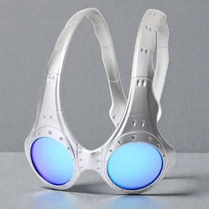 May include: Silver-coloured goggles with blue lenses and a clear, curved head strap. The goggles have a futuristic design with visible rivets and a streamlined shape. The lenses reflect a bright blue colour, and the overall aesthetic is modern and sporty.