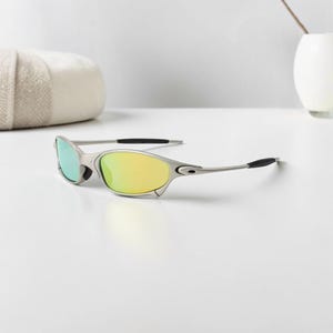 May include: Silver-framed sunglasses with yellow and green mirrored lenses. The sunglasses have a sleek, modern design with black temple tips. The lenses reflect a gradient of colors, adding a stylish touch to the eyewear.