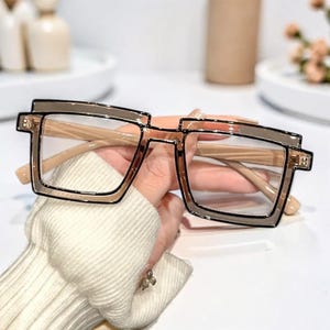 Könnte beinhalten: Quadratisch gerahmte Brille mit hellbraunem und schwarzem Rahmen. Die Brille hat ein doppellagiges Rahmendesign und klare Gläser. Die Bügel der Brille sind hellbraun. Die Brille wird von einer Person gehalten, die einen weißen Pullover trägt.