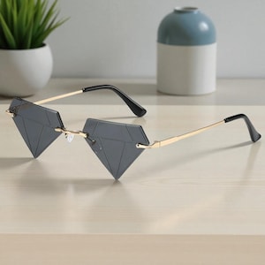 May include: Diamond-shaped sunglasses with a gold-coloured frame and dark grey lenses. The sunglasses have a unique geometric design and black temples. The glasses are displayed on a light-coloured surface.
