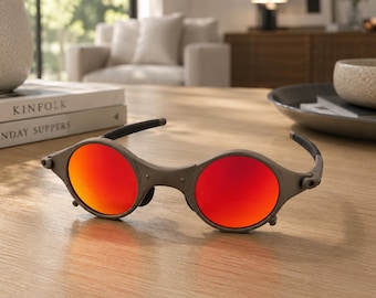 Peut inclure: Lunettes de soleil avec une monture grise et des verres ronds rouge-orange vif. Les lunettes sont posées sur une table en bois. Les branches des lunettes sont noires.