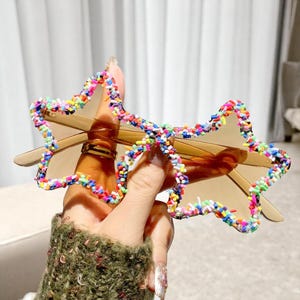 K&ouml;nnte beinhalten: Sternf&ouml;rmige Sonnenbrille mit braunem Rahmen und get&ouml;nten Gl&auml;sern. Die Sternkonturen sind mit bunten Streuseln verziert. Die Sonnenbrille wird von einer Hand gehalten.