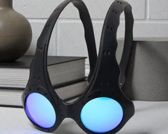 Peut inclure: Lunettes noires avec des verres bleus. Les lunettes ont un design unique avec une monture incurvée et une sangle. Les verres ronds reflètent une teinte bleue. Les lunettes sont posées sur une surface blanche.