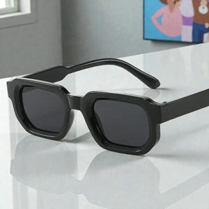 May include: Black square-framed sunglasses with dark lenses. The sunglasses have a thick, angular frame and a classic, modern design. The lenses are dark, providing shade and protection from the sun.