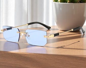 May include: Rimless sunglasses with light blue lenses and gold-colored frames. The temples are black and gold. The sunglasses are resting on a wooden surface, with a white pot and a cactus in the background.