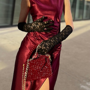 Può includere: Una donna indossa un lungo abito di raso bordeaux con uno spacco laterale. Indossa guanti di pizzo neri e porta una piccola borsa a mano di perline rosse con una catena dorata.