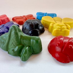 Puede incluir: Un conjunto de nueve crayones de cera de colores con forma de insectos y mariposas. Los crayones incluyen una mariquita roja, un saltamontes verde, una mariquita negra, una abeja amarilla, una mariposa azul y tres mariposas rojas, moradas y naranjas.