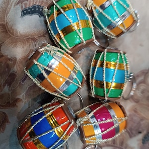 May include: A collection of six miniature drum-shaped ornaments. Each drum features a colorful design with bands of orange, blue, green, and red, accented with gold and silver trim. The drums have silver ring attachments.