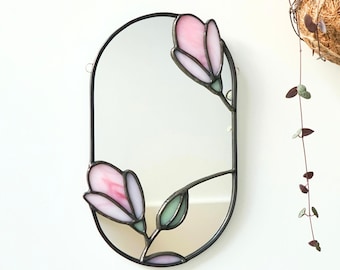 May include: An oval wall mirror framed in dark metal, featuring stained glass accents in the shape of two pink flower buds and a small green leaf. The floral elements are integrated into the mirror's edge, creating a decorative border.