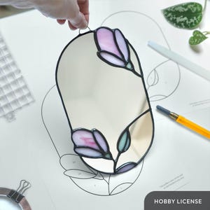 Op de afbeelding: Een ovaalvormige glas-in-lood spiegel met een zwarte rand en bloemmotieven. De bloemen zijn roze en wit met groene bladeren. De spiegel wordt door een hand vastgehouden. De tekst "HOBBY LICENSE" is zichtbaar.