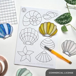 Op de afbeelding: Een sjabloon voor glas-in-lood met genummerde secties voor een schelp, een zeester en een nautilus. Het vel is omringd door afgewerkte glas-in-loodstukken, waaronder een blauwe schelp, een gele schelp, een witte schelp en een groene schelp. De tekst "COMMERCIAL LICENSE" staat onderaan het vel.