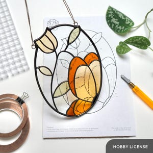 Op de afbeelding: Gebrandschilderd glazen vlinderkunstwerk met oranje en gele vleugels, omlijst in zwart metaal. Het kunstwerk hangt aan een bruin touwtje. Een patroon en knutselspullen staan op de achtergrond. De tekst "HOBBY LICENSE" staat onderaan.