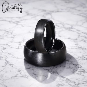 May include: Two black rings with a brushed metal finish are shown. One ring stands upright, the other lies flat. The rings are set against a marble background. The words "Glentify Handmade" are in the upper left corner.