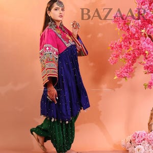 Puede incluir: Una mujer con un atuendo tradicional con una parte superior rosa, un vestido azul y pantalones verdes. El atuendo presenta intrincados bordados y adornos. La imagen incluye el texto "KOCHA BAZAAR" y est&aacute; ambientada sobre un fondo melocot&oacute;n con flores rosas.