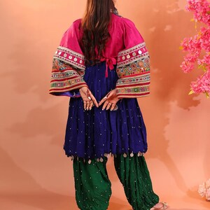 Puede incluir: Un conjunto vibrante con una chaqueta rosa con mangas bordadas y adornos plateados. El atuendo incluye una blusa azul fluida y pantalones verdes fruncidos. La vista trasera muestra los intrincados detalles del dise&ntilde;o tradicional.