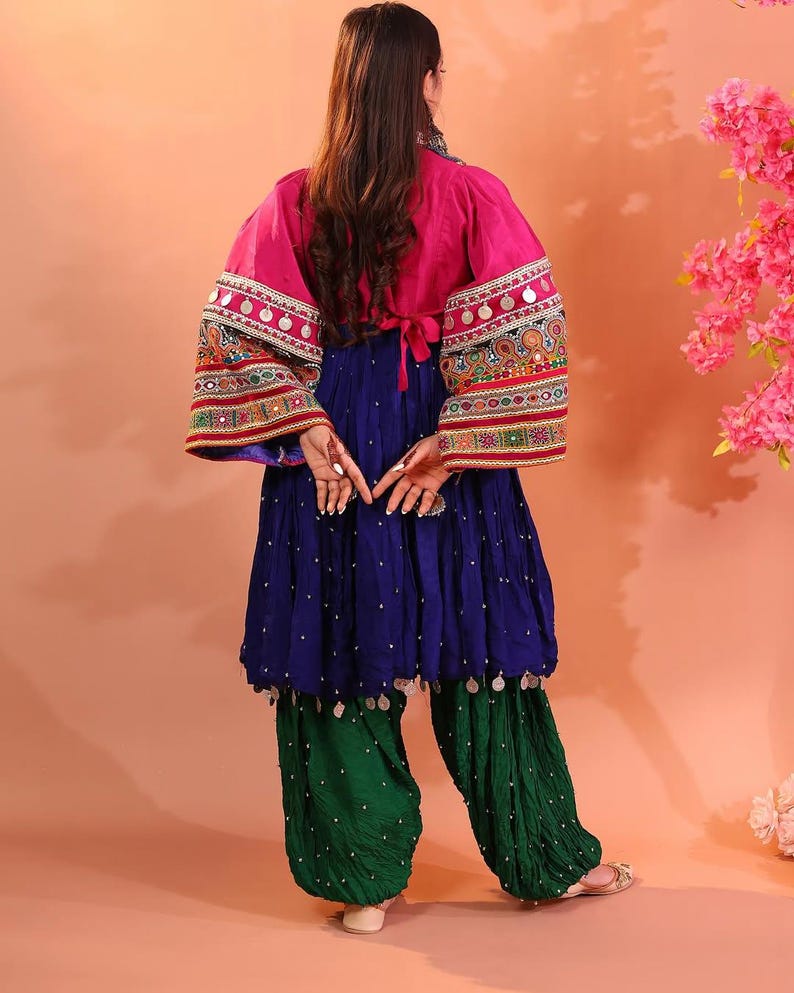 Puede incluir: Un conjunto vibrante con una chaqueta rosa con mangas bordadas y adornos plateados. El atuendo incluye una blusa azul fluida y pantalones verdes fruncidos. La vista trasera muestra los intrincados detalles del dise&ntilde;o tradicional.